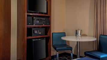 A hotel room with a flat-screen TV, a minibar, a small refrigerator, a round table with a chair, and a bed.