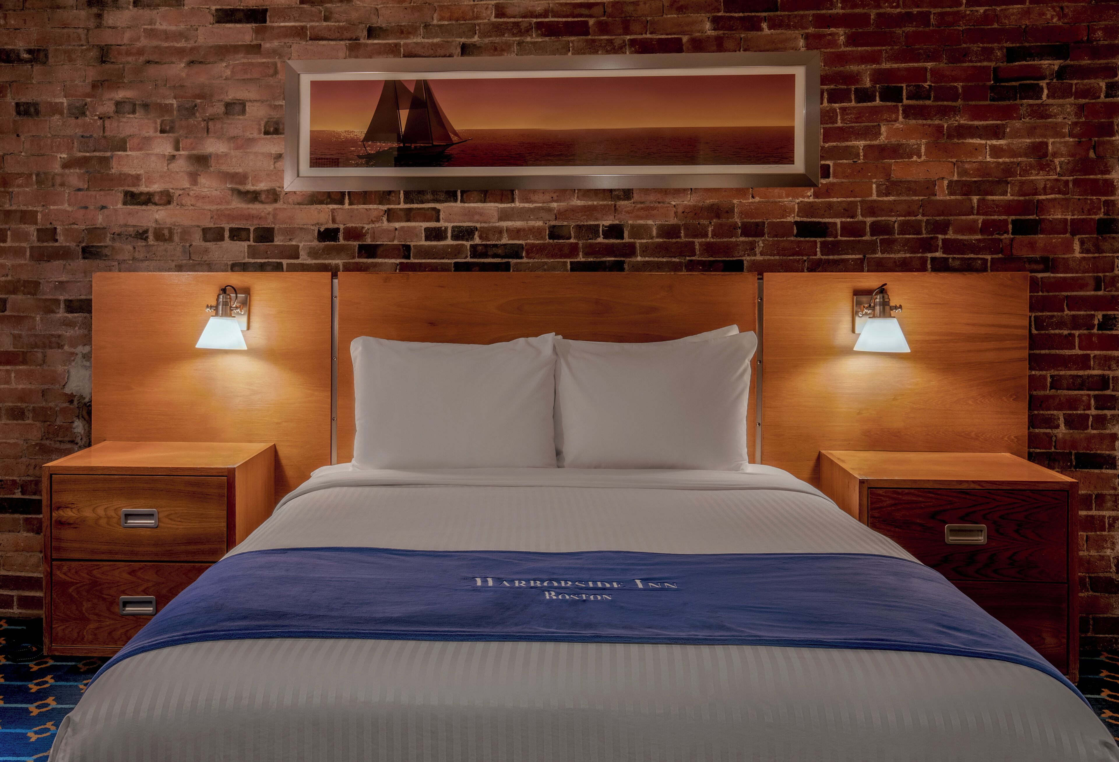 Habitación de hotel con pared de ladrillo, una cama con ropa blanca, dos mesitas de noche con lámparas y un cuadro enmarcado sobre la cama.