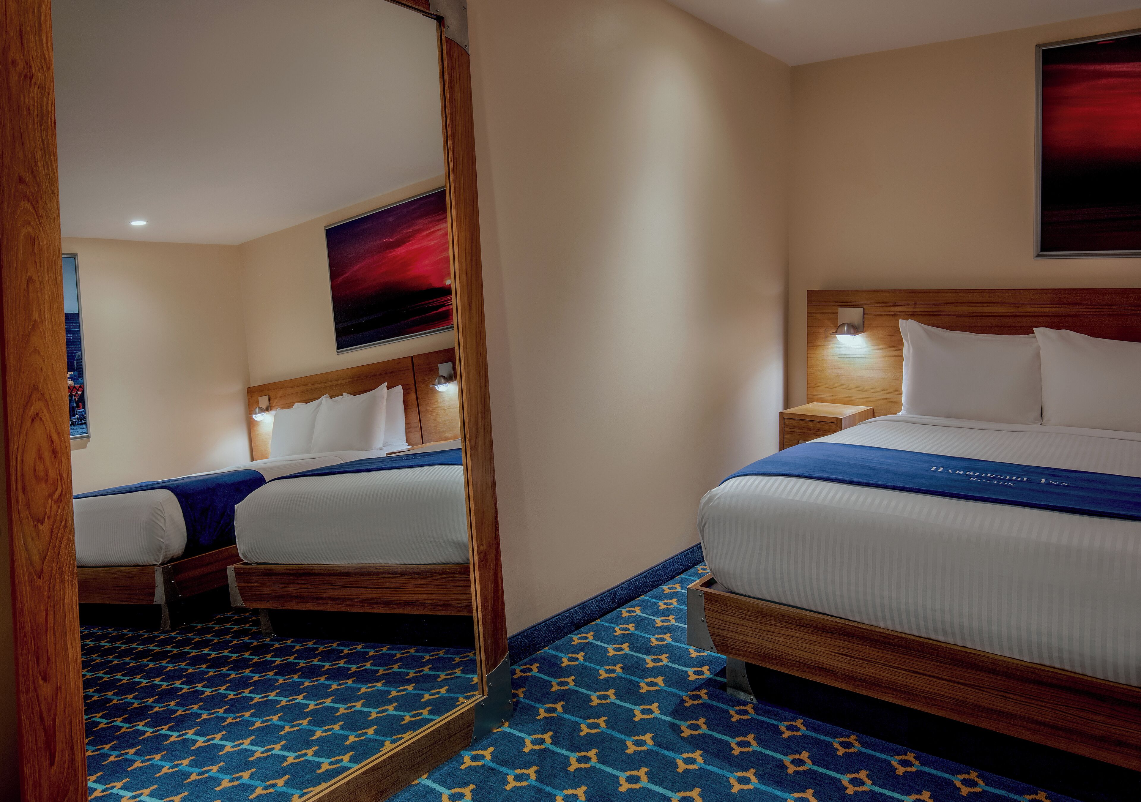A hotel room with two beds, a mirror, a nightstand, a wall-mounted lamp, and a large painting.