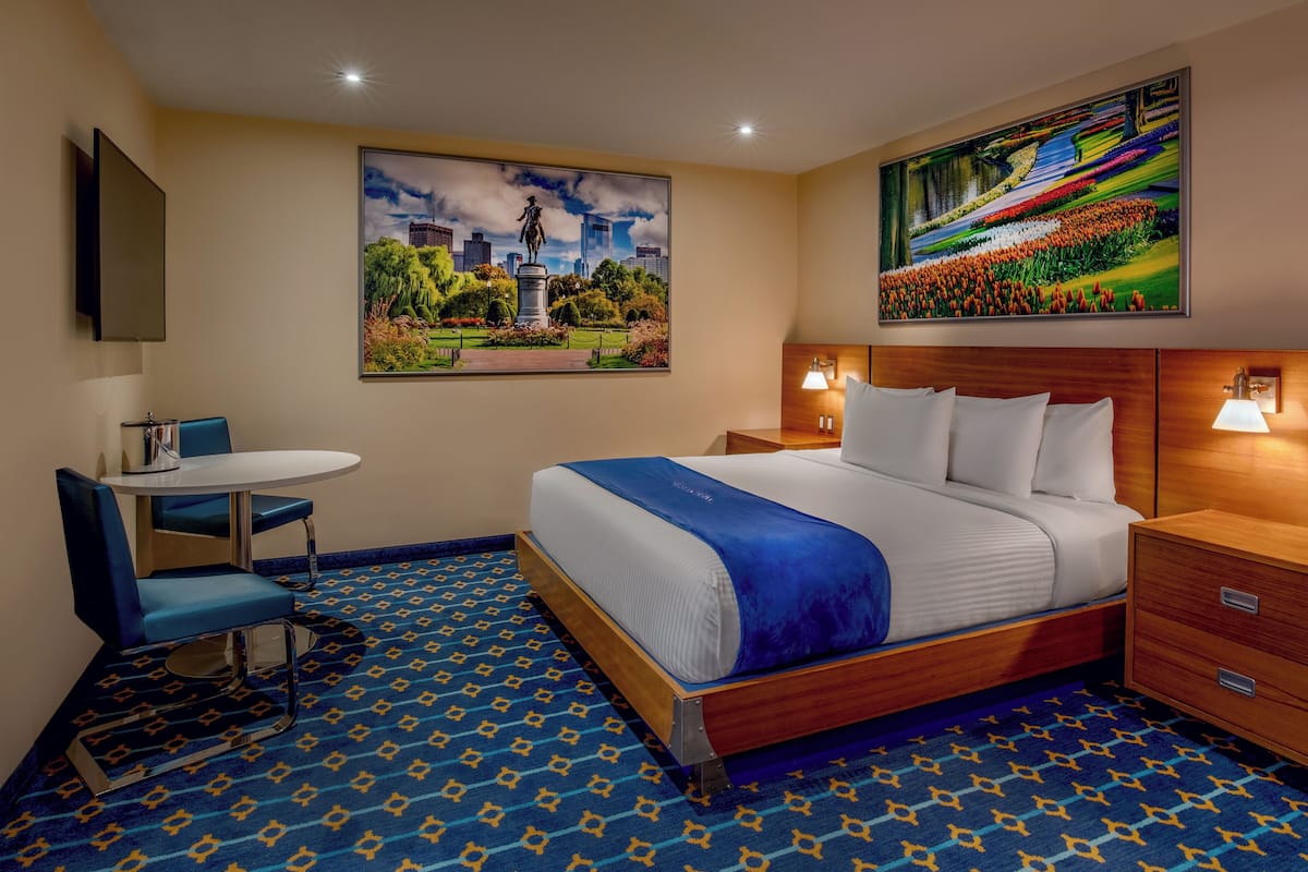 A hotel room with a bed, bedside tables, a desk, and chairs. There are two paintings on the walls.