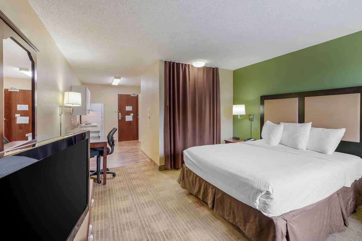 Ein Hotelzimmer mit einem großen Bett, einem Schreibtisch mit Stuhl, einem Spiegel und einem fensterbepflanzten Fenster.