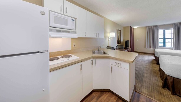 A compact hotel room with a kitchenette including a microwave, sink, and cabinets. There is a bed and a window with curtains. - Extended Stay America Suites Orange County Huntington Beach (Huntington Beach)