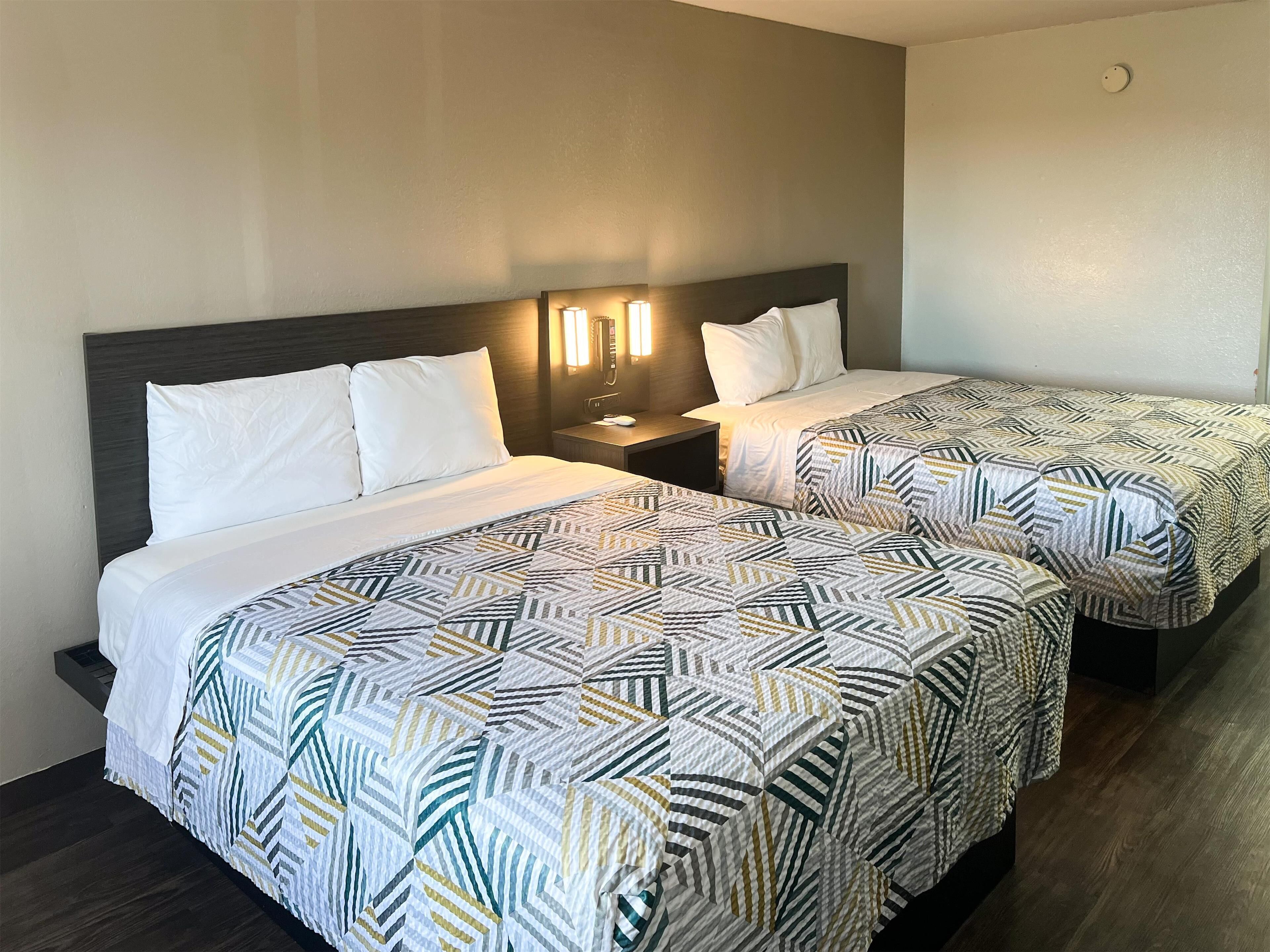 A hotel room with two beds, each with a patterned bedspread, a nightstand with a lamp, and a wall-mounted light fixture.