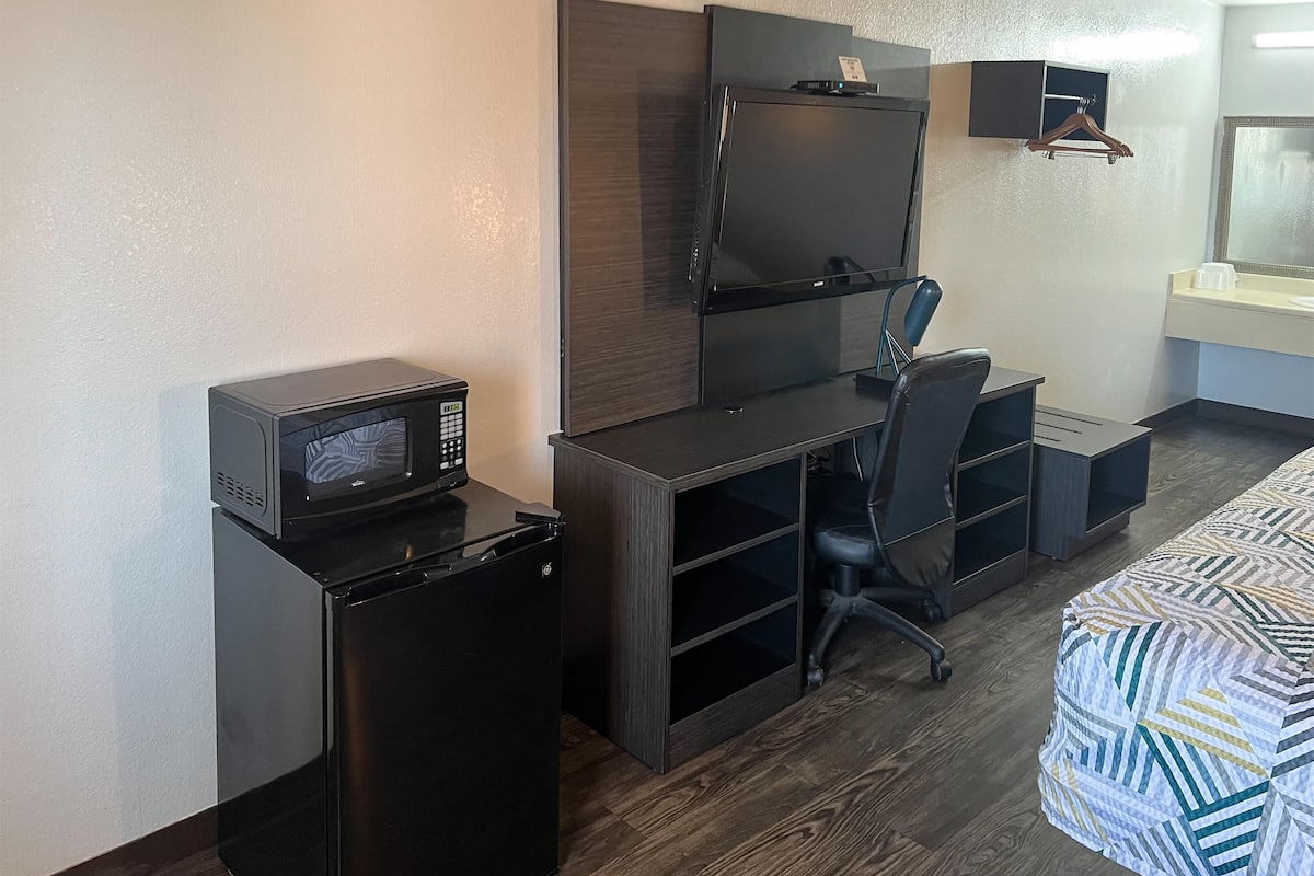 Quarto de hotel compacto com cozinha compacta, uma mesa com cadeira, cama e televisão fixada na parede.