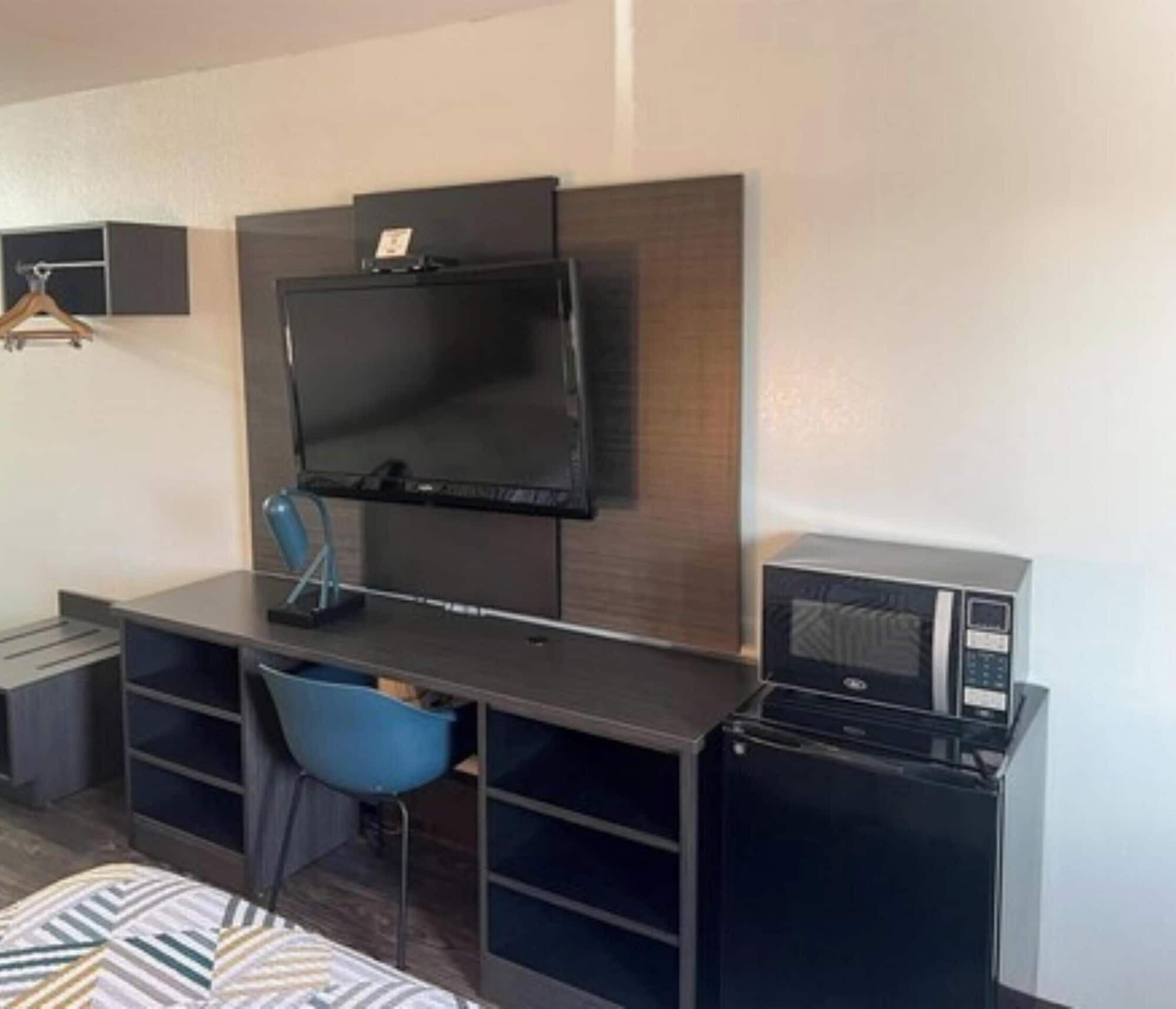 A compact hotel room with a flat-screen TV mounted on the wall, a small desk with a chair, and a microwave on the right.