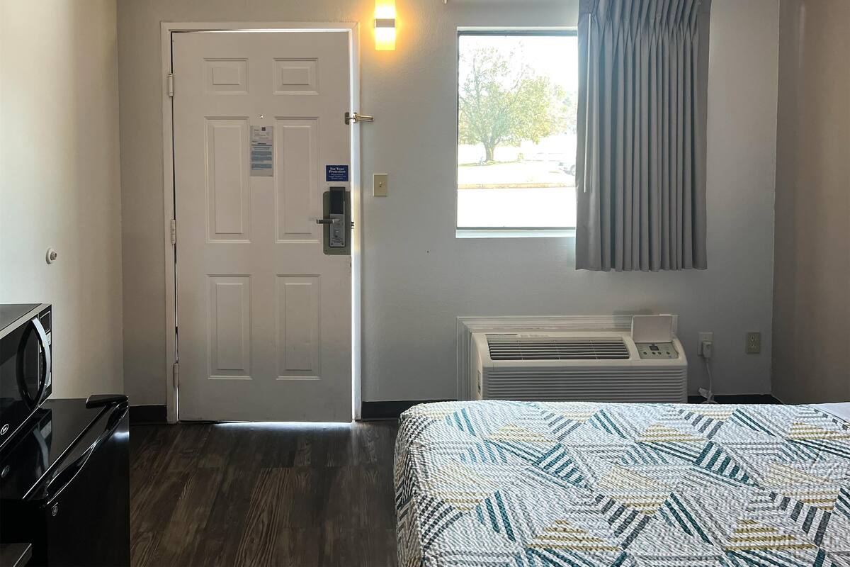Quarto de hotel com cama, escrivaninha, porta com dispositivo de segurança, janela com cortinas e ar-condicionado fixado na parede.