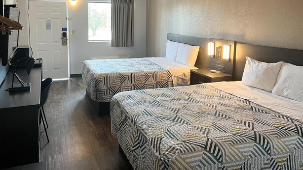 Habitación de hotel con dos camas, un escritorio y una silla. Se observa una puerta con un cartel, una ventana con cortinas y una lámpara fijada a la pared.