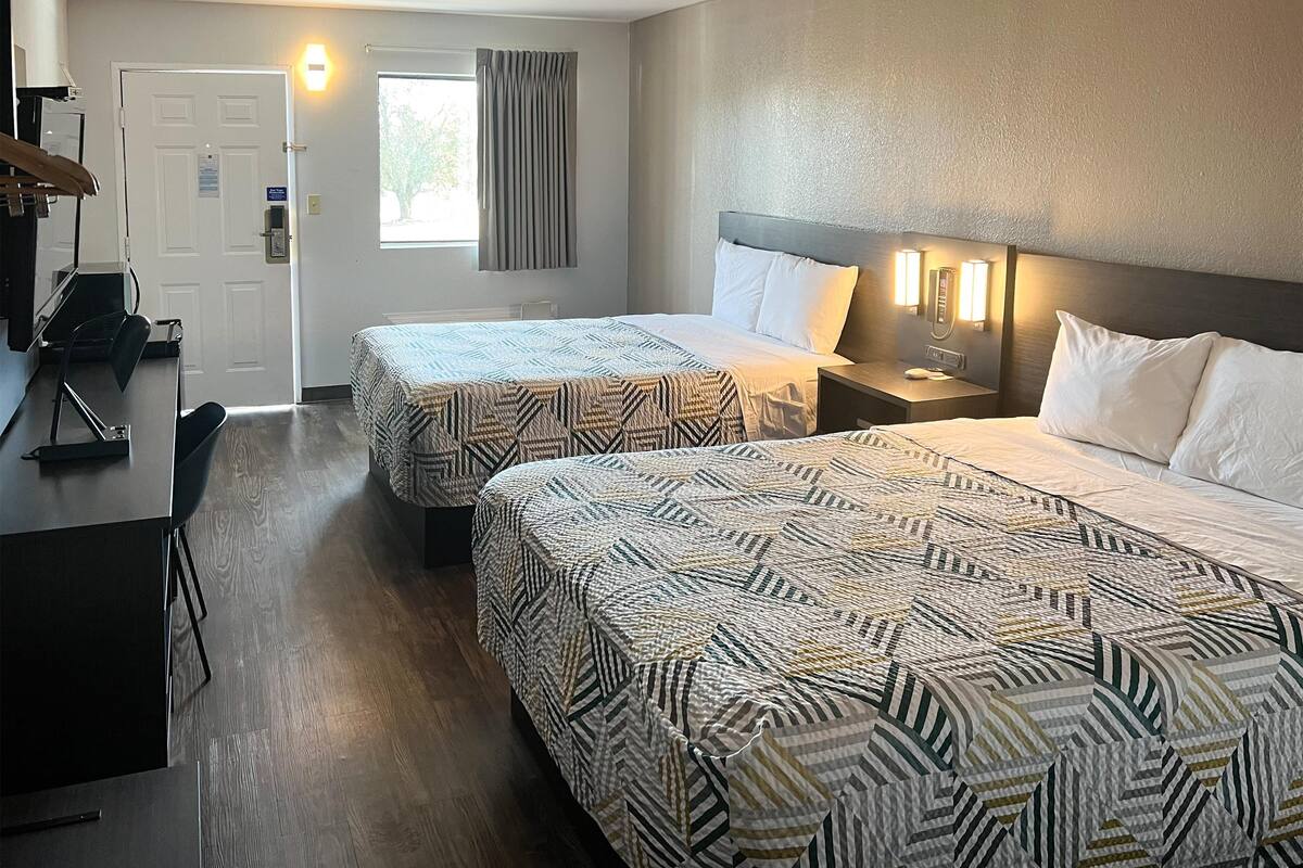 Quarto de hotel com duas camas, uma escrivaninha e uma cadeira. Há uma porta com sinalização, uma janela com cortinas e um ponto de luz fixado na parede.