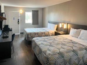 A hotel room with two beds, a desk, and a chair. There is a door with a sign, a window with curtains, and a wall-mounted light fixture.