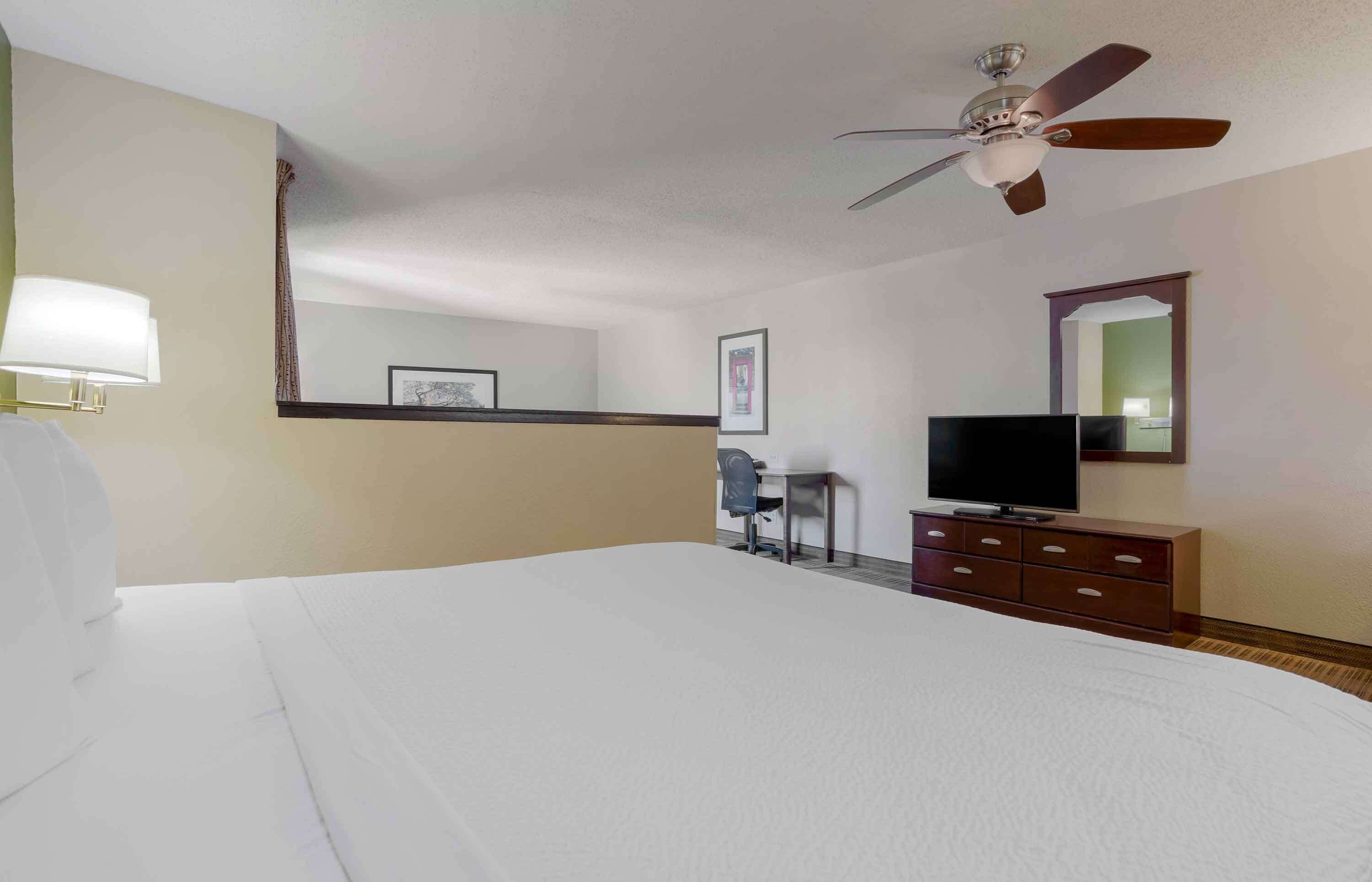 A hotel room with a large bed, a desk with a chair, a television, a dresser, and a ceiling fan.