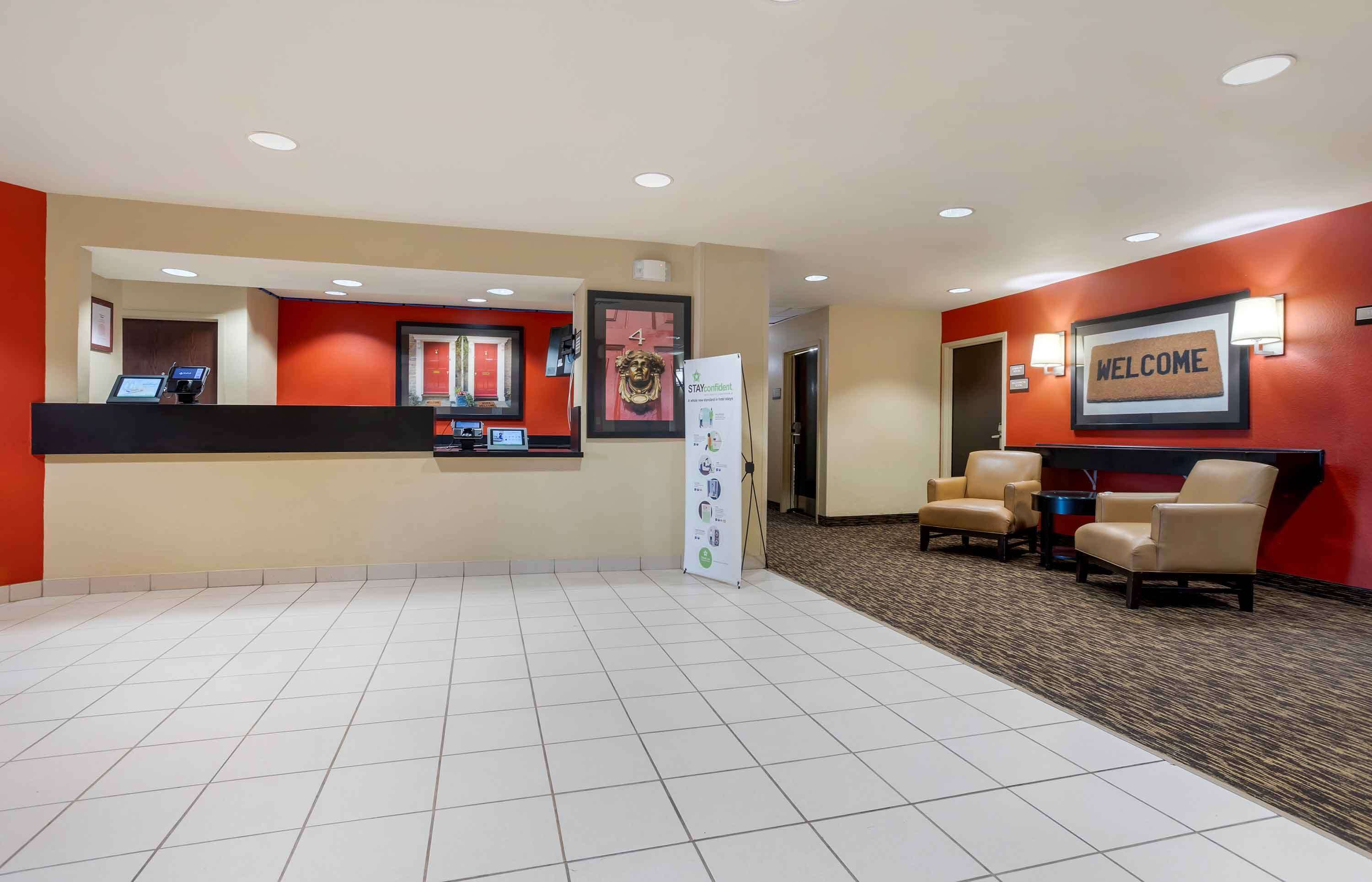 A hotel reception area with a reception desk, a 'WELCOME' sign, a 'STAYCONFIDENT' sign, and a seating area with chairs and a small table.