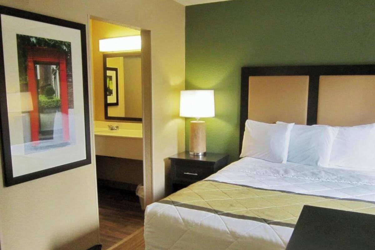 A hotel room with a bed, a nightstand with a lamp, a framed picture on the wall, and a bathroom visible through an open door.