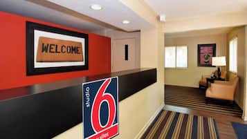 A hotel reception area with a 'WELCOME' sign, a '6 suites' sign, a reception desk, a door, a lounge area with a sofa and a lamp, and a striped carpet.