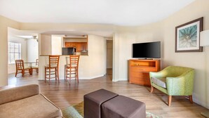 A living room with a sofa, ottoman, and a television on a stand. There is a dining area with chairs and a table.