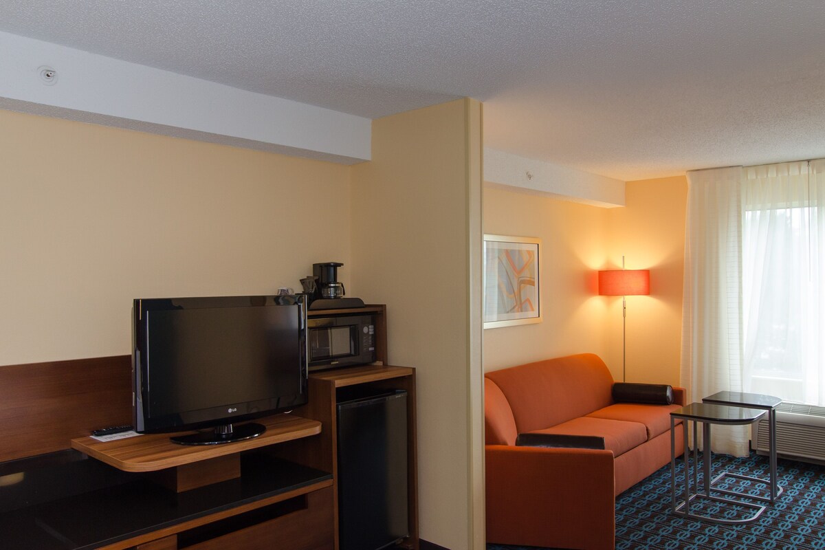 Hotelkamer met flatscreen-tv, magnetron, kleine koelkast, oranje bank, bijzettafeltje en raam met gordijnen.