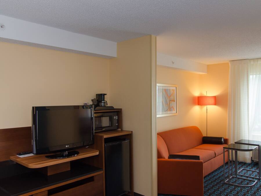 Quarto de hotel com televisão de tela plana, micro-ondas, mini-geladeira, sofá laranja, mesa lateral e janela com cortinas.