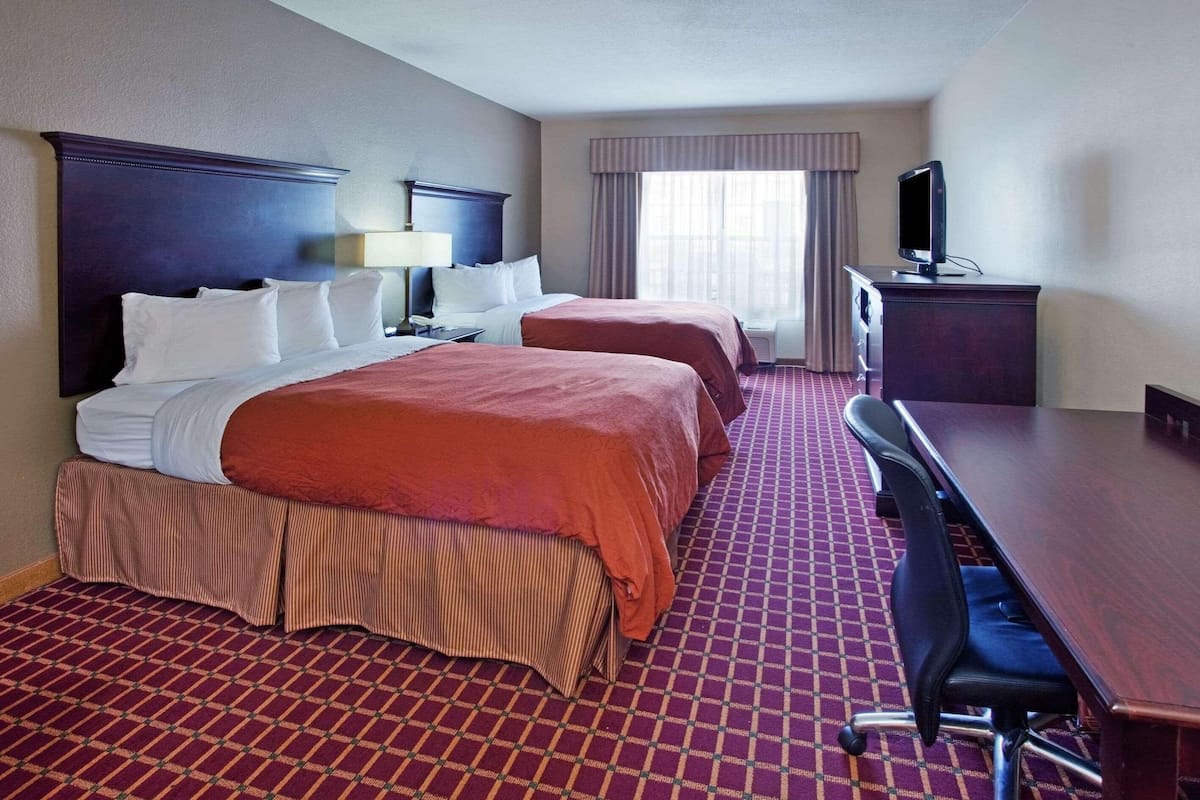 A hotel room with two beds, a desk, a television, and a window with curtains.
