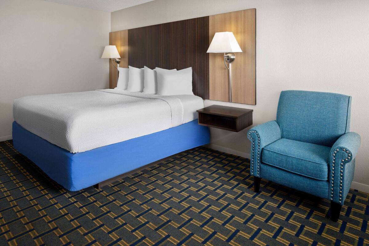 Une chambre d’hôtel avec un lit, un fauteuil bleu, une table de chevet et deux lampes fixées au mur.