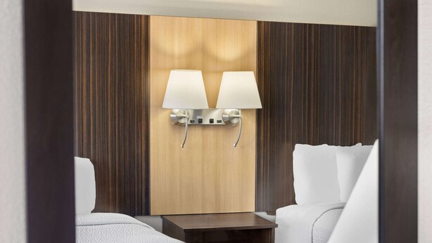 A hotel room with a wooden panel wall, two bedside lamps, a small wooden nightstand, and two white beds. - Days Inn by Wyndham Tonawanda/Buffalo (Tonawanda)