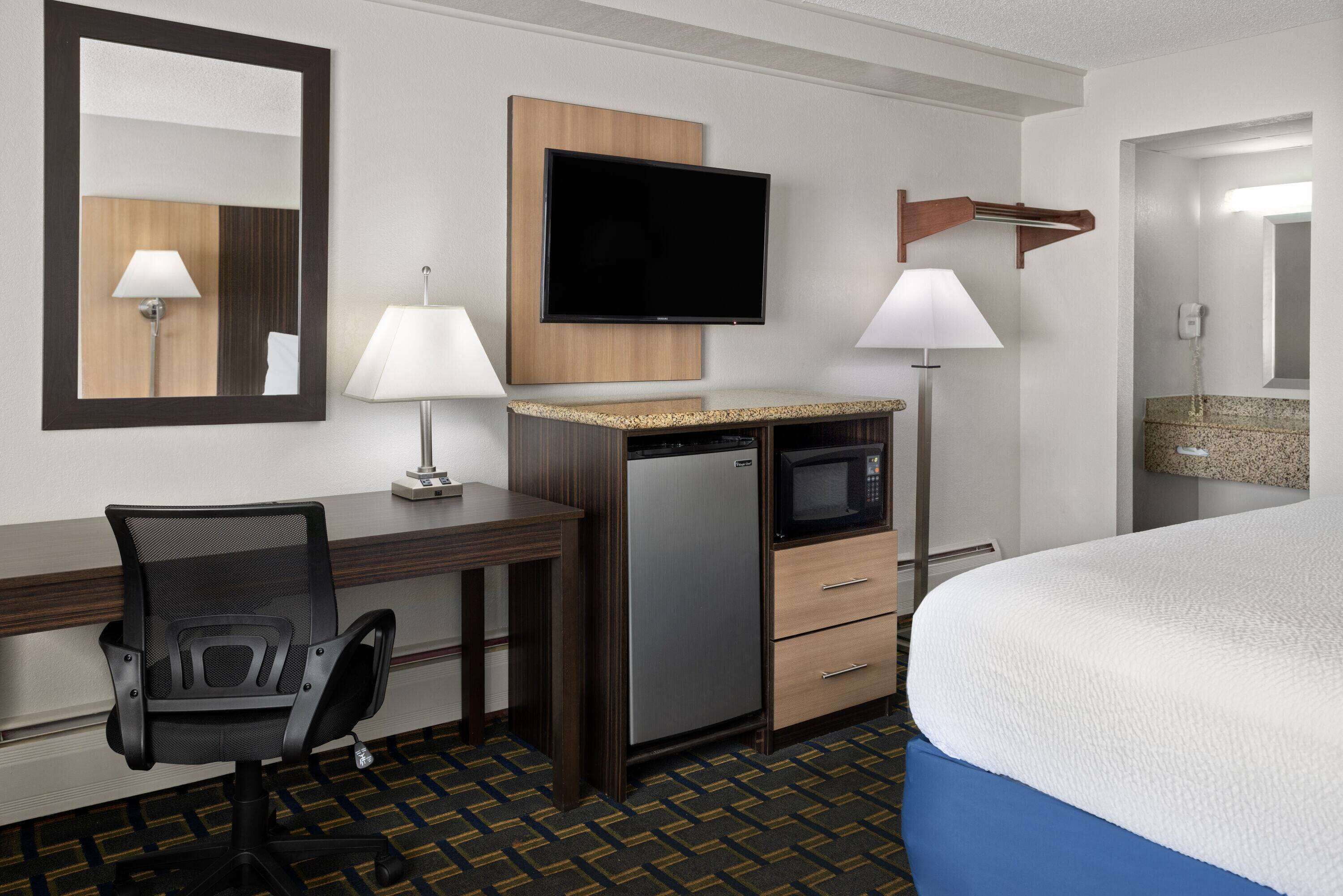 A hotel room with a bed, desk, chair, TV, microwave, and a small refrigerator.