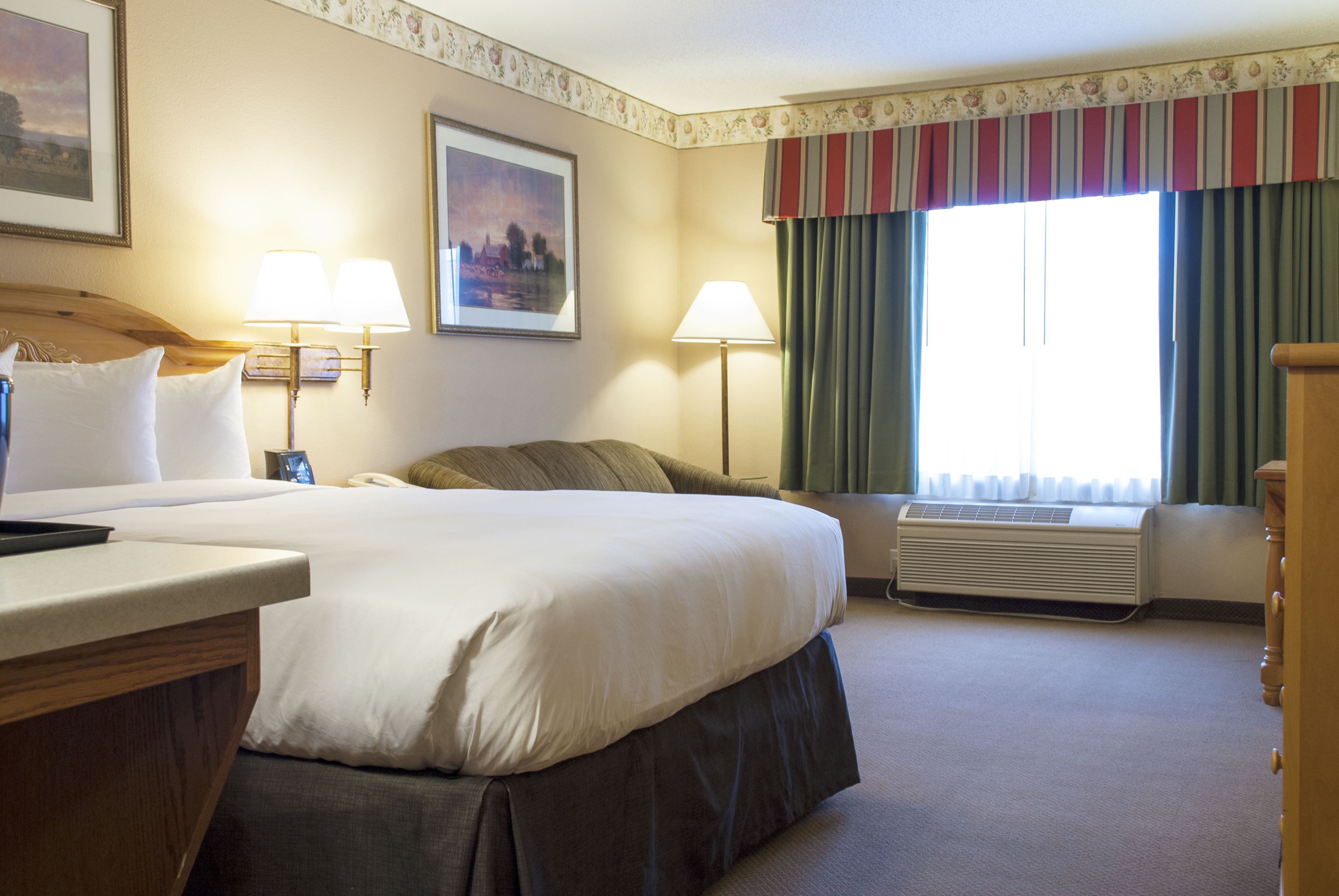 A hotel room with a large bed, two bedside lamps, a window with curtains, a wall-mounted air conditioner, and a wooden dresser.