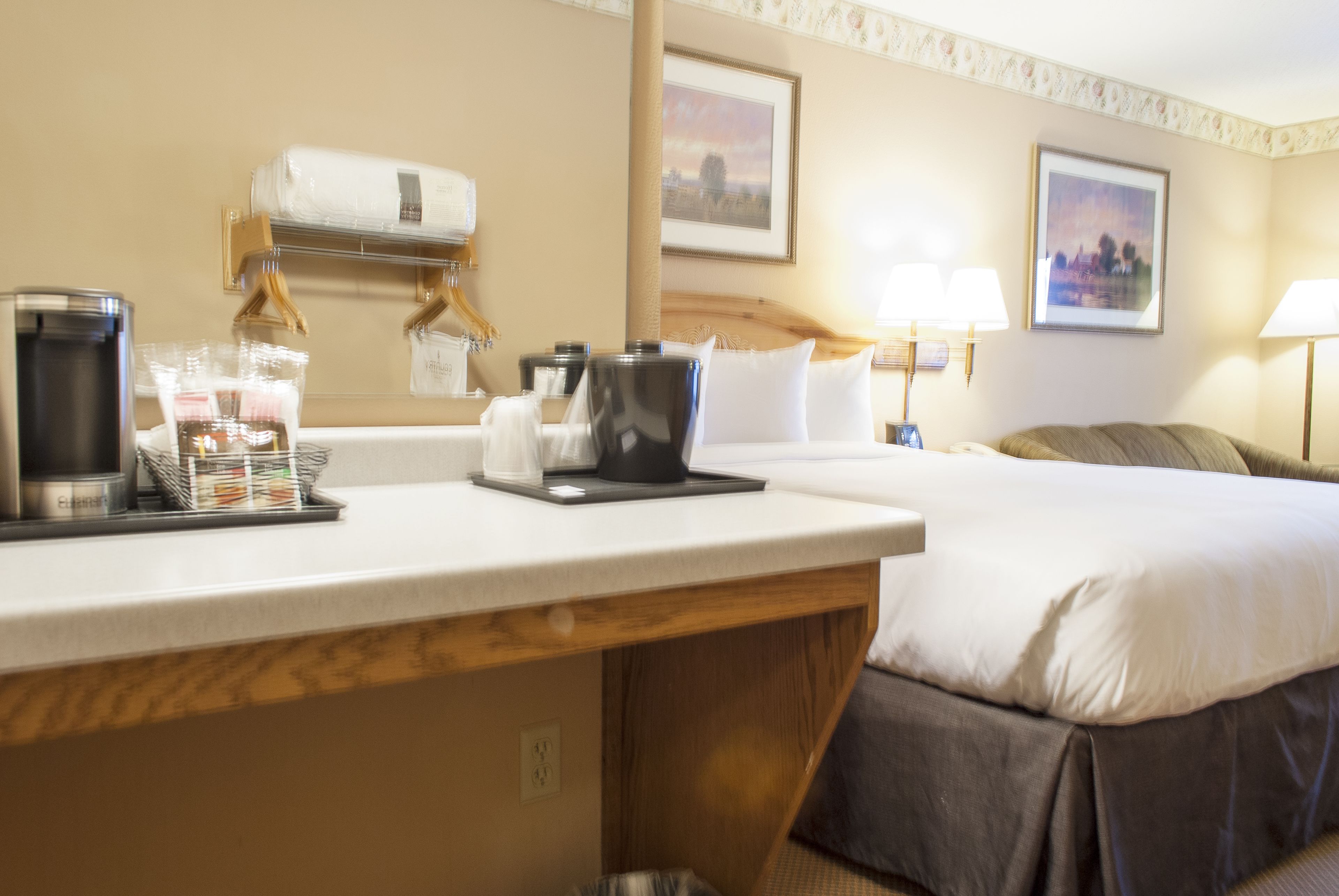 A hotel room with a bed, a desk with a coffee maker, a lamp, and a picture on the wall.