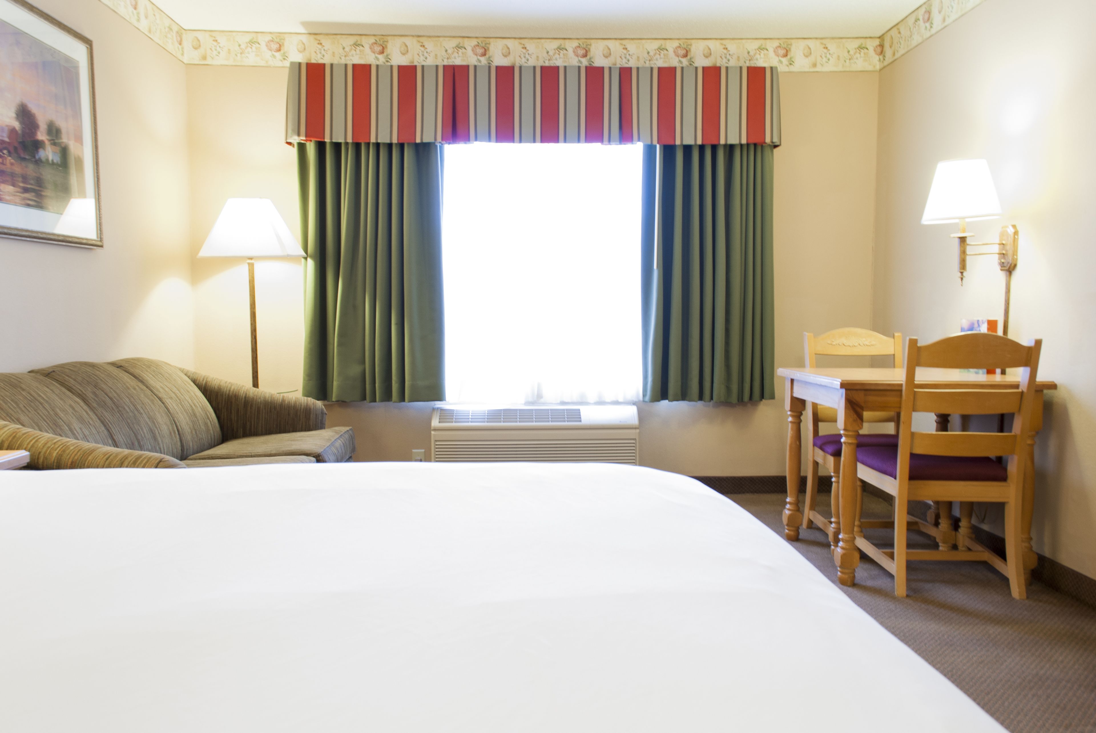 Quarto de hotel com uma cama grande, uma escrivaninha com cadeira, uma janela com cortinas e duas luminárias.