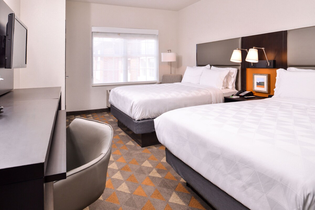 Une chambre d’hôtel avec deux lits, un bureau, une chaise, une fenêtre et une lampe.