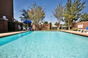 A rectangular swimming pool with clear blue water, surrounded by brick walls and trees. There are lounge chairs and a blue umbrella near the pool.