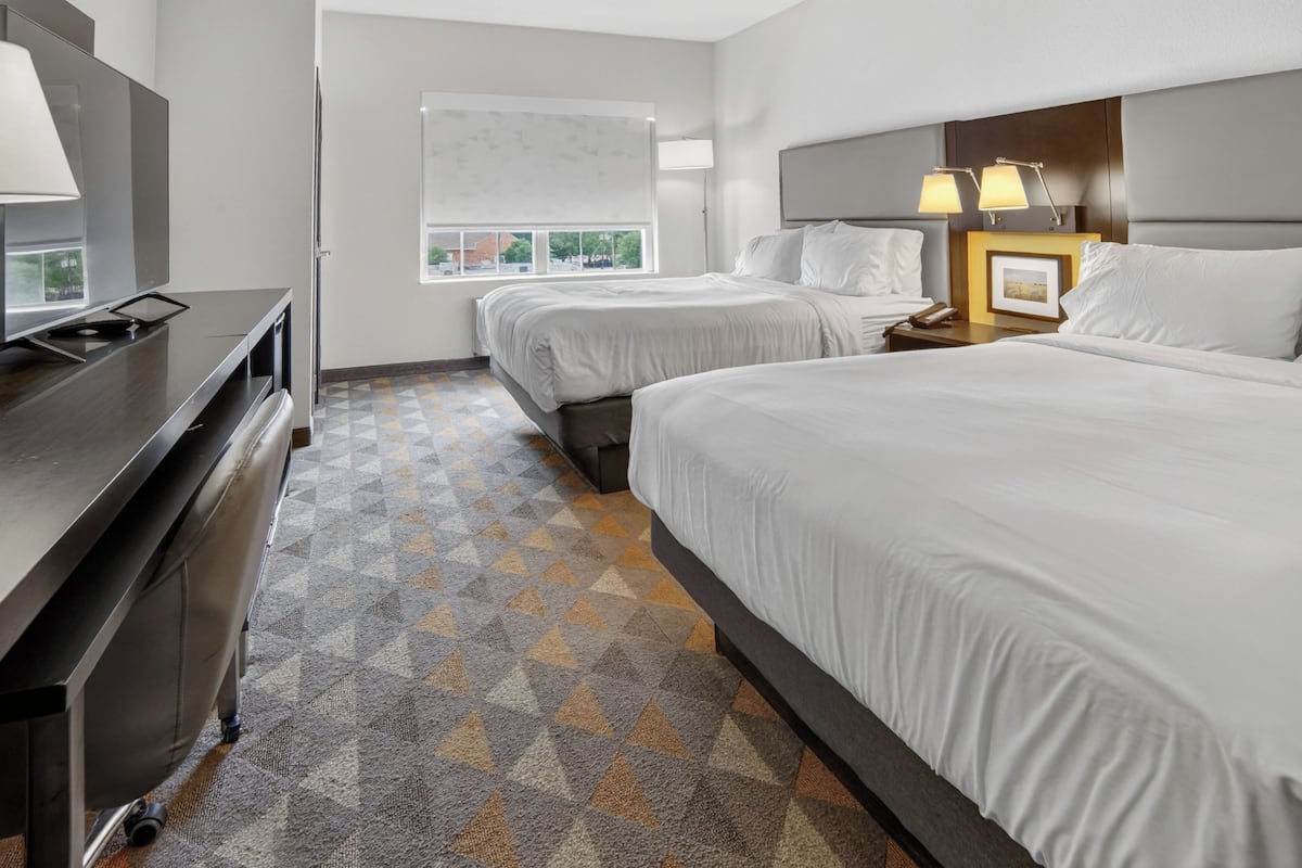 A hotel room with two beds, a desk with a TV, a window with a view, and a patterned carpet.