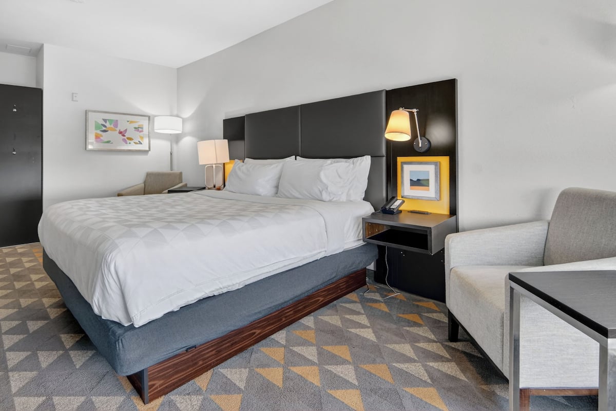 A hotel room with a bed, bedside tables, a lamp, a chair, and a wall-mounted picture frame.