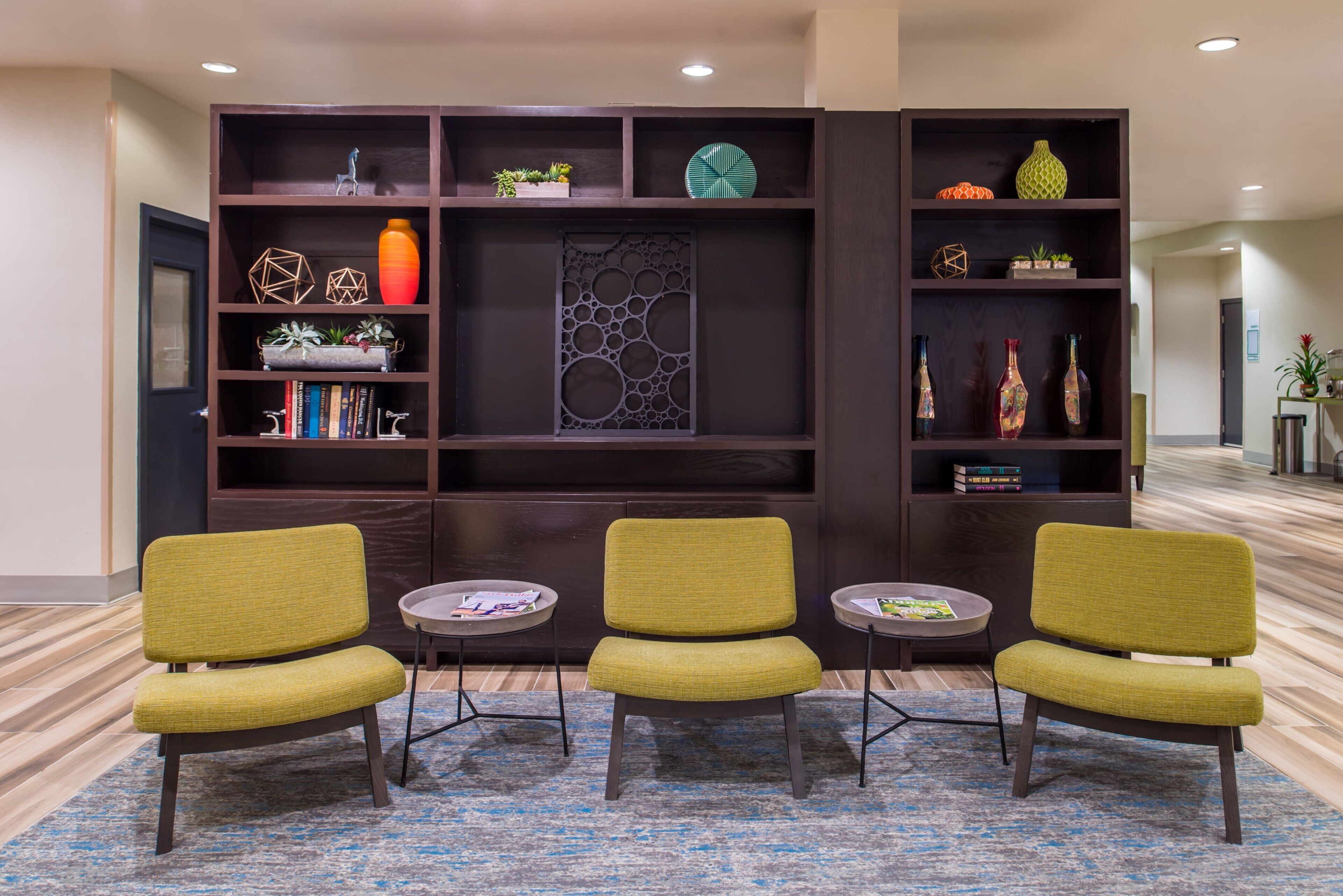 A lobby area with a dark wood bookshelf, three green upholstered chairs, and a patterned rug.