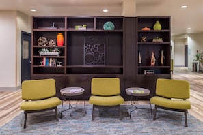 A lobby area with a dark wood bookshelf, three green upholstered chairs, and a patterned rug.
