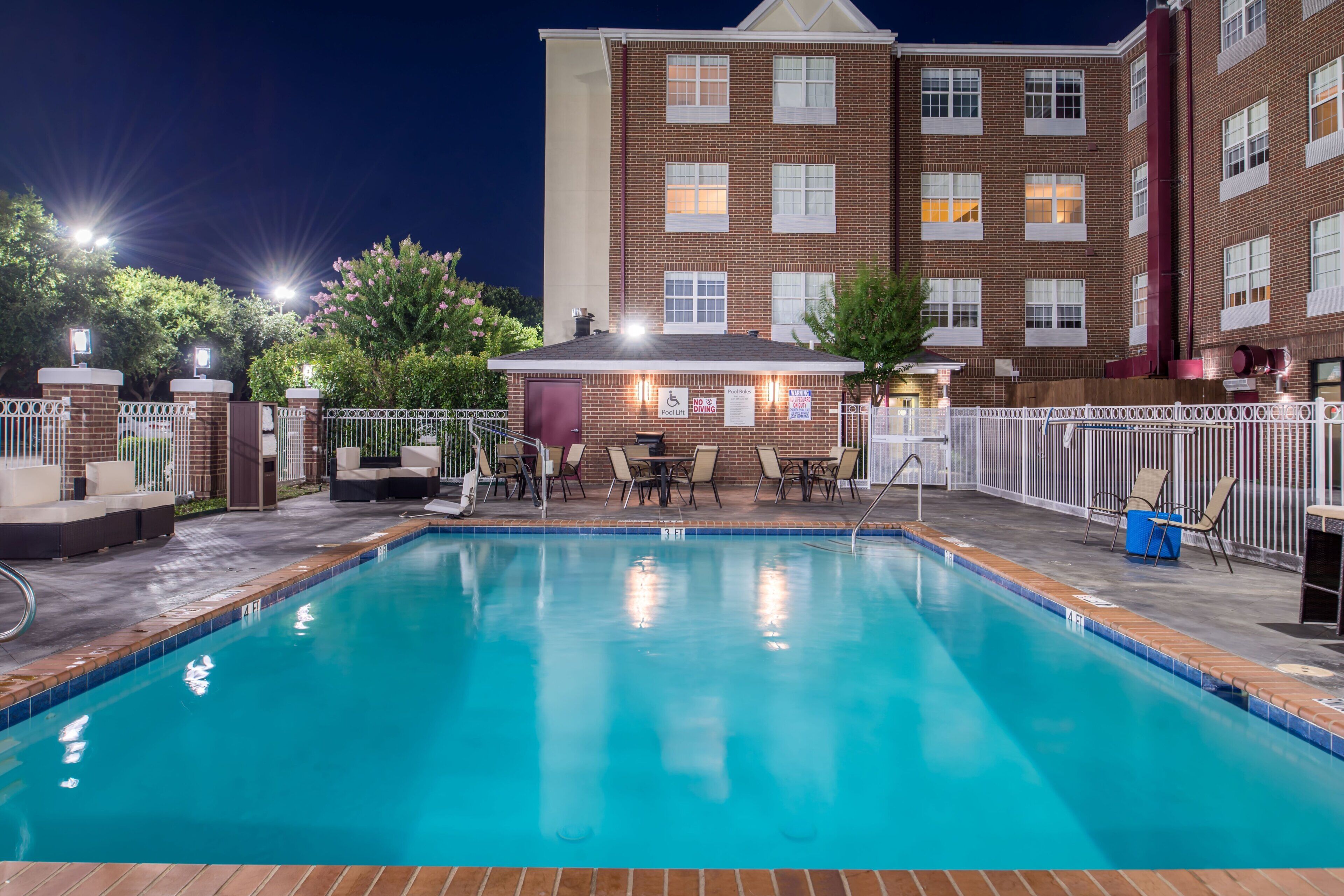 A swimming pool with a sunken hot tub, surrounded by a deck with chairs and tables, and a brick building in the background.