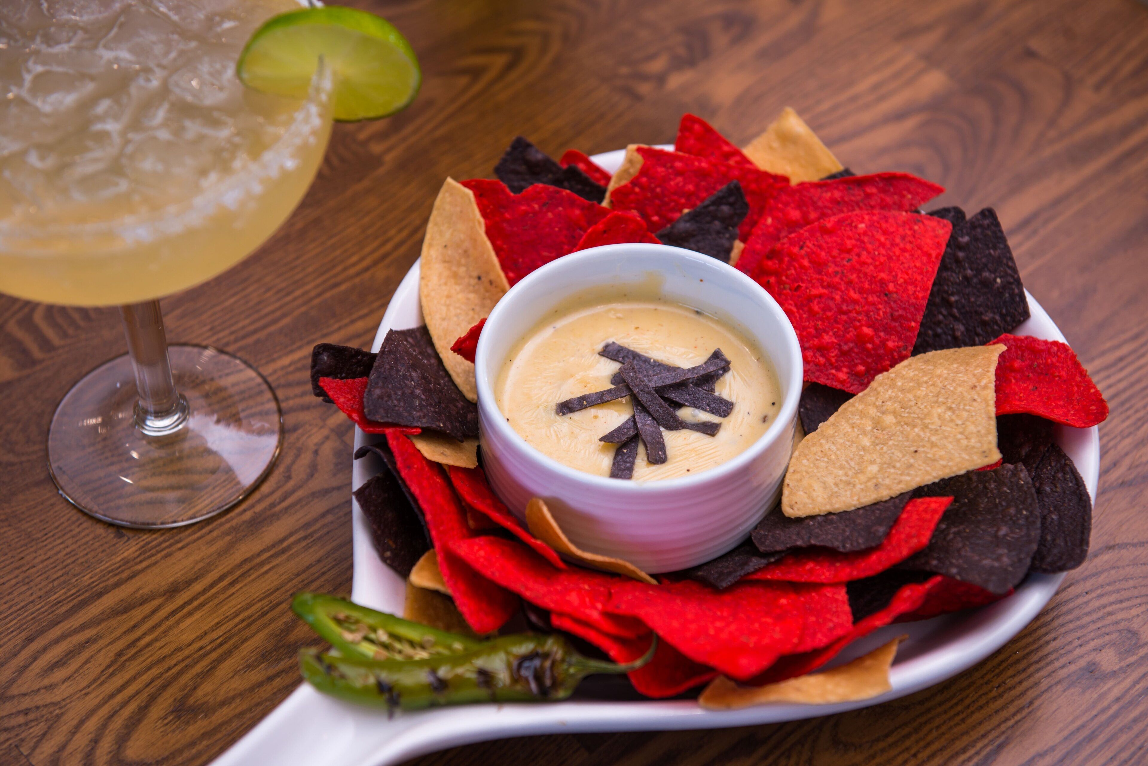 A bowl of dip surrounded by tortilla chips on a plate, with a glass of a yellowish beverage and a lime wedge in the background.