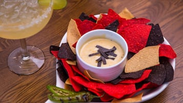 A bowl of dip surrounded by tortilla chips on a plate, with a glass of a yellowish beverage and a lime wedge in the background.