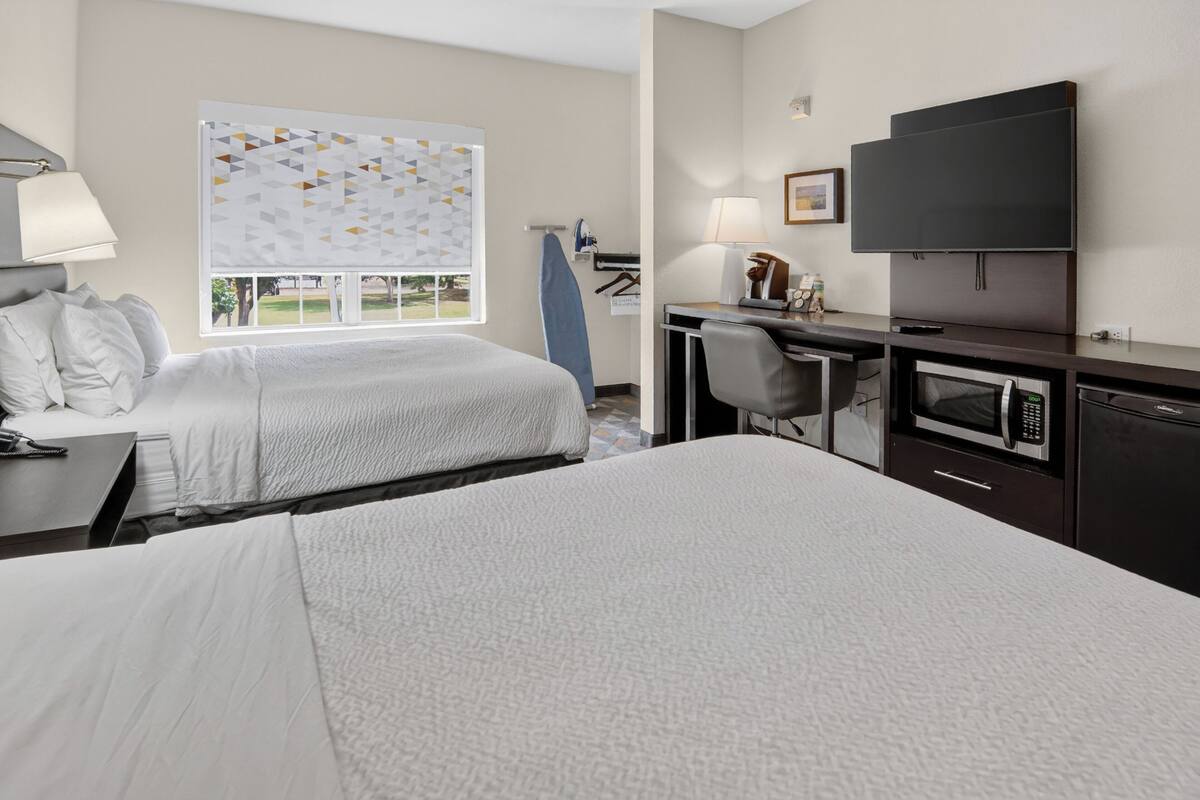 A hotel room with two beds, a desk with a chair, a flat-screen TV, a microwave, and a window with a view of the outdoors.