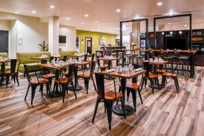 A hotel dining area with multiple tables and chairs, a reception desk, and a green sofa. - Holiday Inn & Suites Dallas-Addison by IHG (Addison)