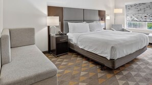 A hotel room with a bed, a sofa, a nightstand, a lamp, and a window with a view. - Holiday Inn & Suites Dallas-Addison by IHG (Addison)
