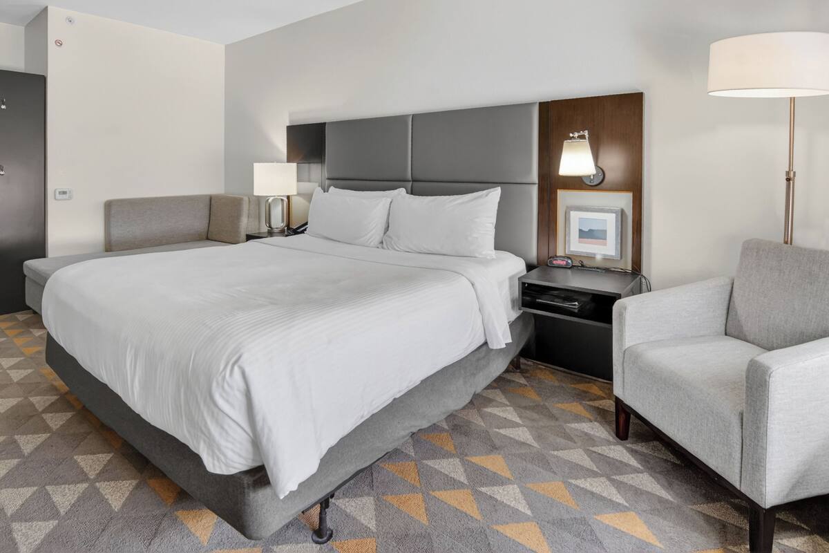 A hotel room with a large bed, a bedside table with a lamp, a chair, and a picture frame on the wall.