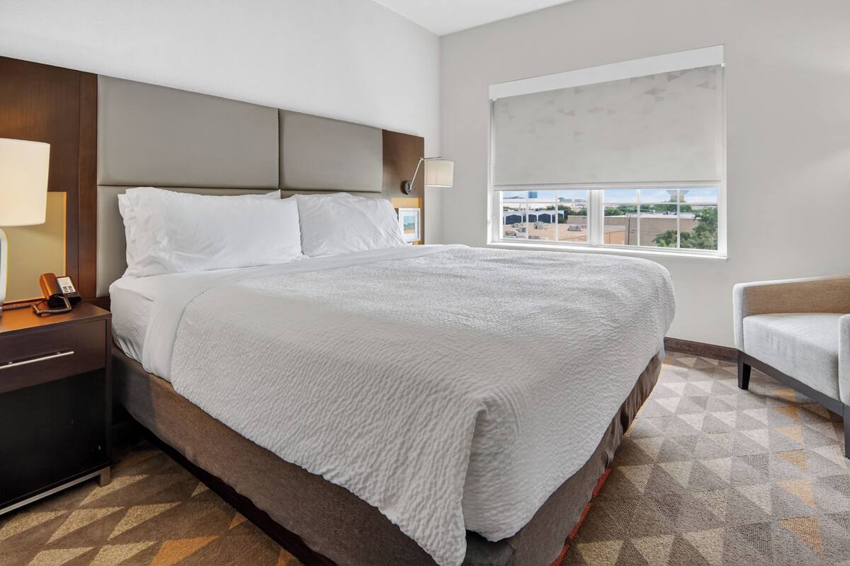 A hotel room with a large bed, a bedside table with a lamp, a chair, and a window with a view.
