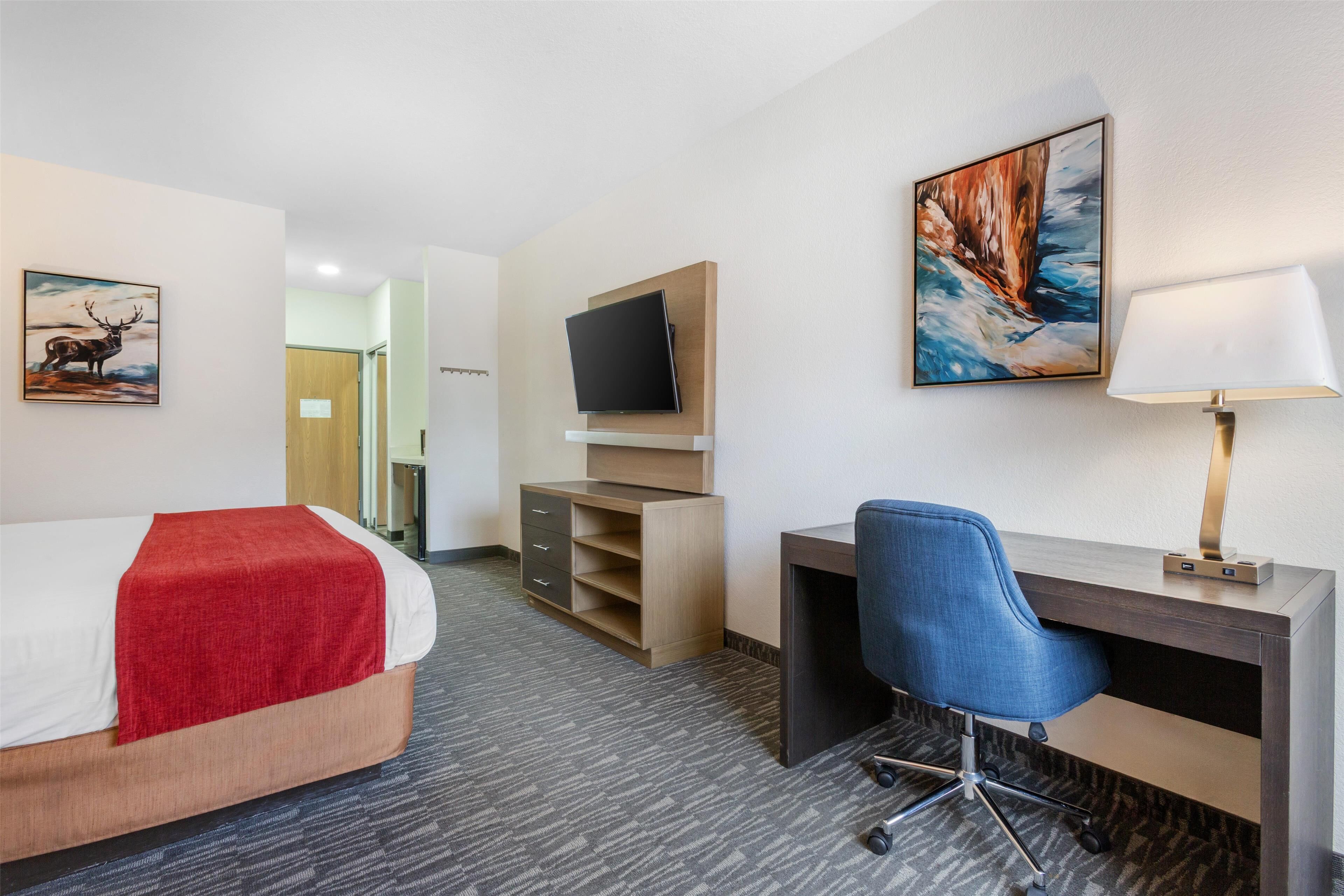A hotel room with a bed, a desk with a chair, a TV mounted on the wall, and artwork on the walls.
