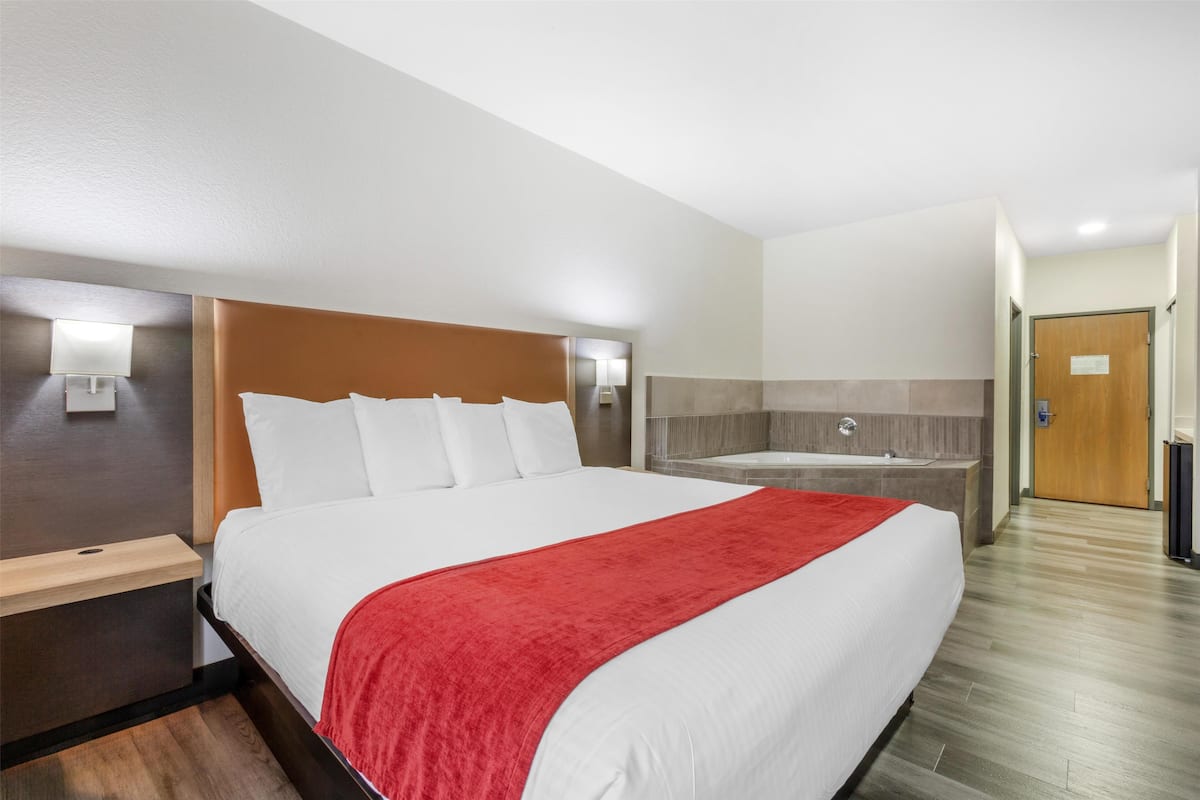 A hotel room with a large bed, a red blanket, a wooden headboard, a nightstand, a wall-mounted lamp, and a bathroom with a bathtub and a shower.