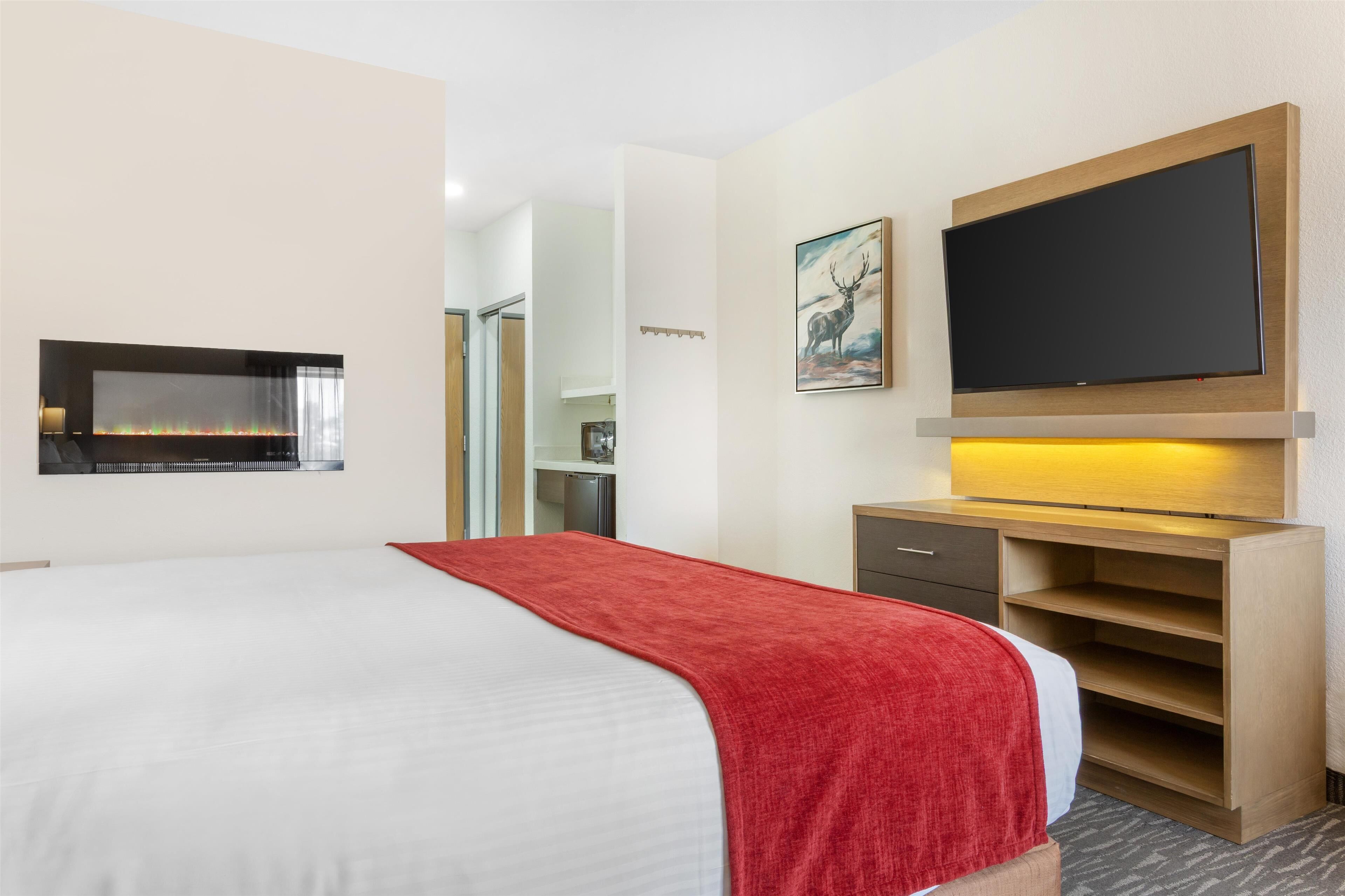 A hotel room with a bed, a TV mounted on the wall, a desk with drawers, and a small shelf.
