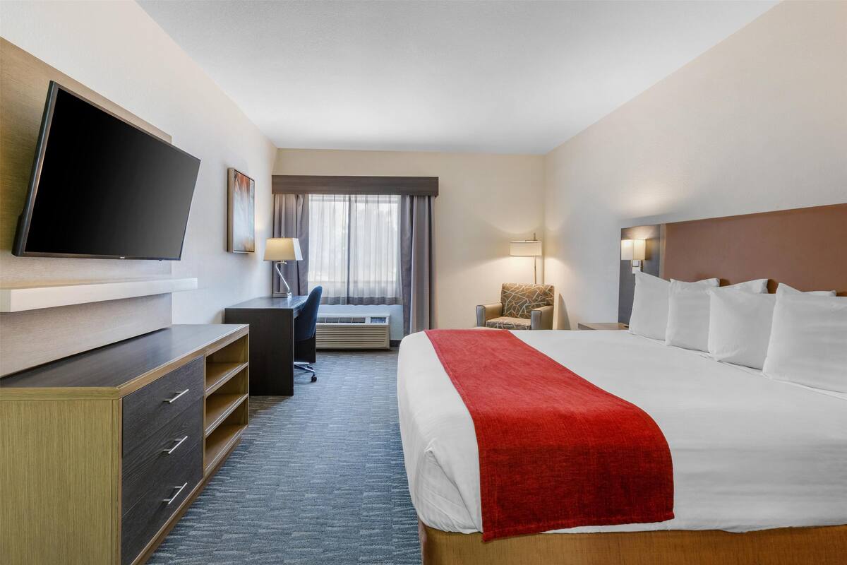A hotel room with a large bed, a flat-screen TV mounted on the wall, a desk with a chair, a window with curtains, and a bedside table with lamps.