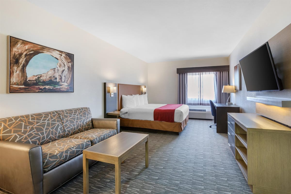 A hotel room with a bed, a sofa, a desk, a TV, and a painting on the wall.