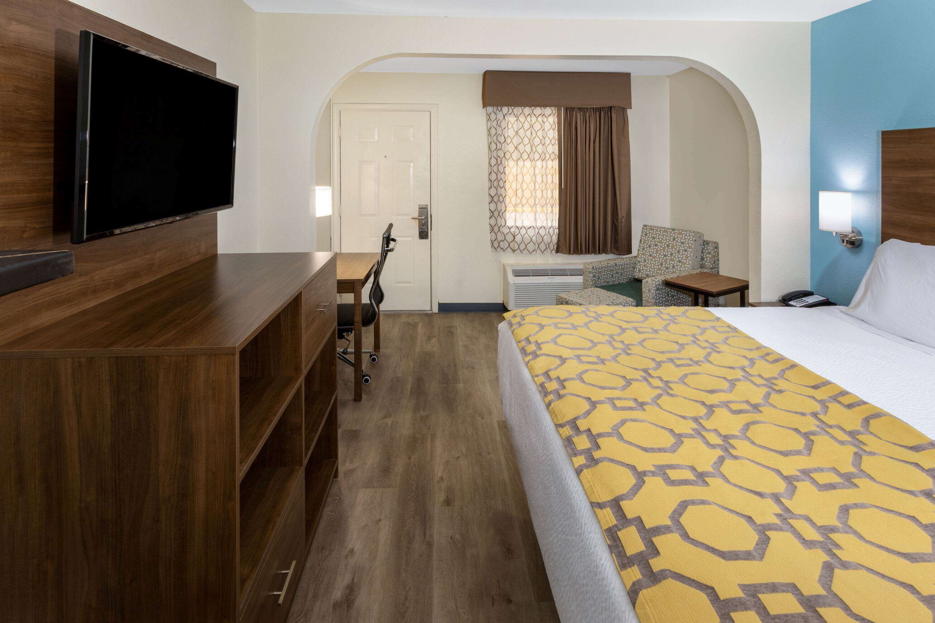 Una habitación de hotel con una cama grande, un televisor montado en un aparador de madera, un escritorio con una silla y un equipo de aire acondicionado.