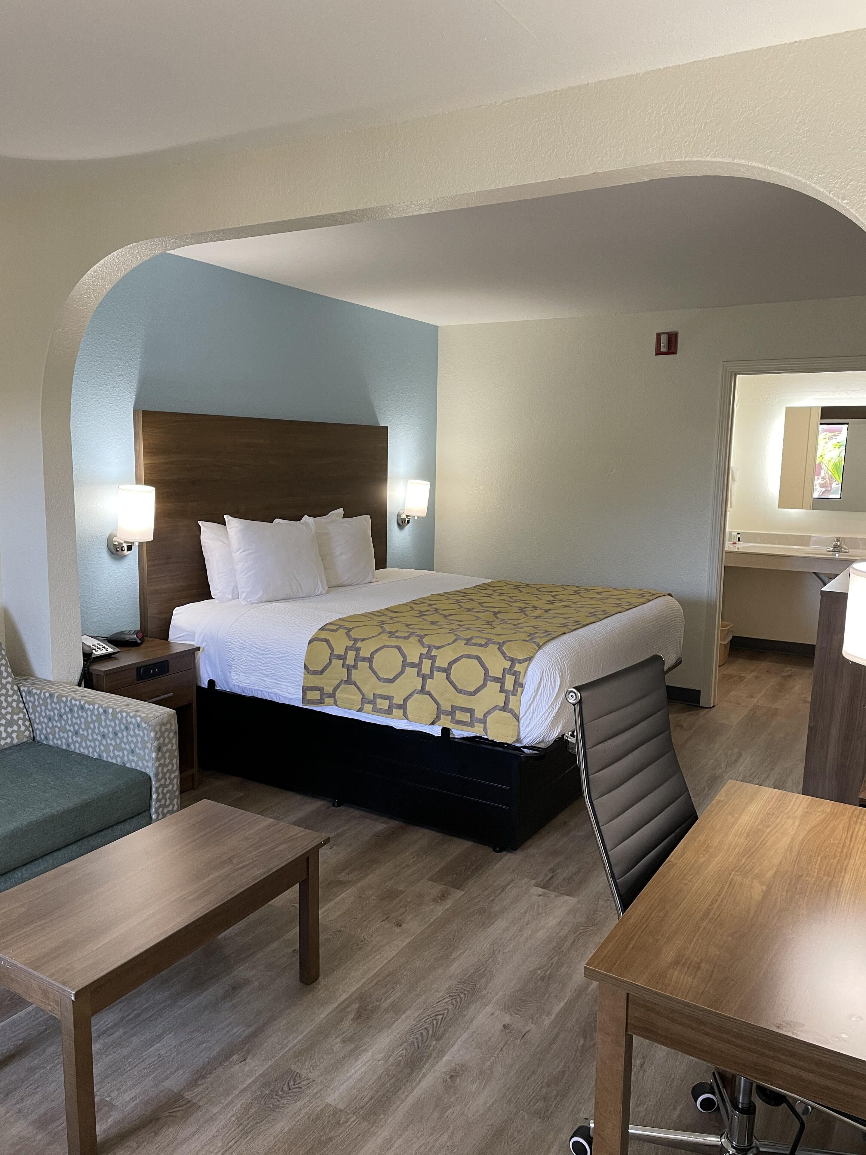 Une chambre d’hôtel comprenant un lit, des tables de chevet, une chaise, un bureau et une salle de bain visible à travers une arche.