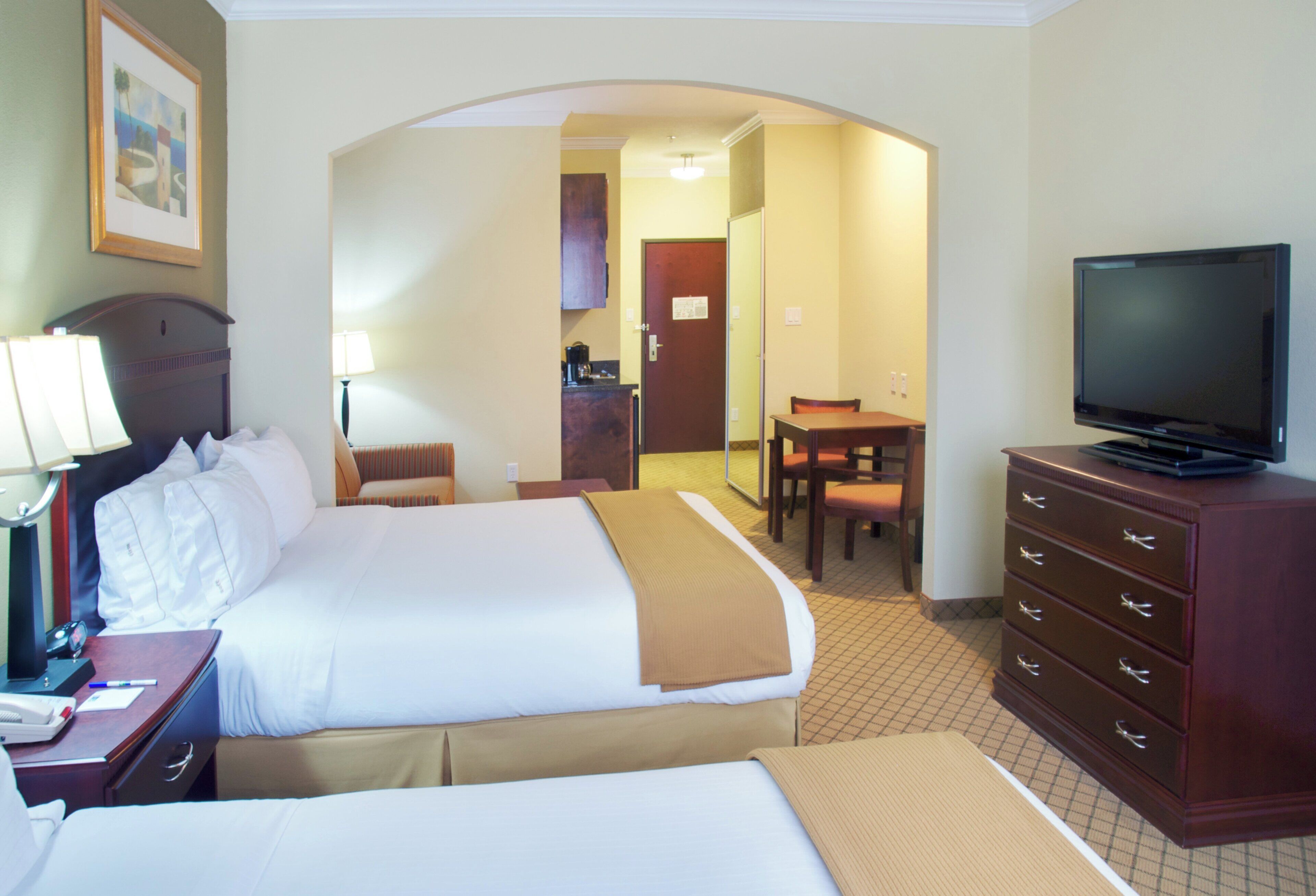 Una habitación de hotel con cama, escritorio con lámpara, televisión y zona de comedor con mesa y sillas.