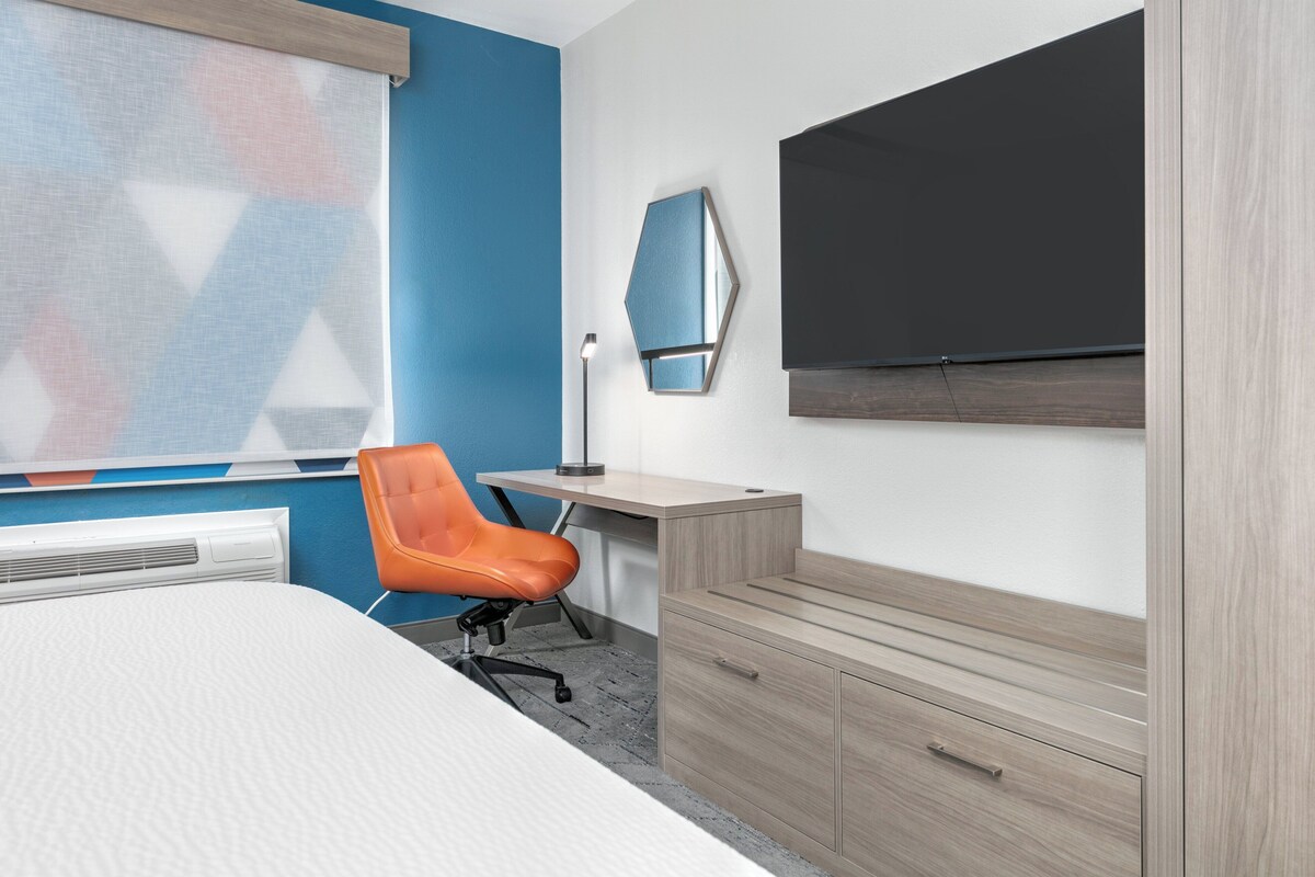 A hotel room with a bed, a desk, an orange chair, a flat-screen TV, a mirror, and a wardrobe.